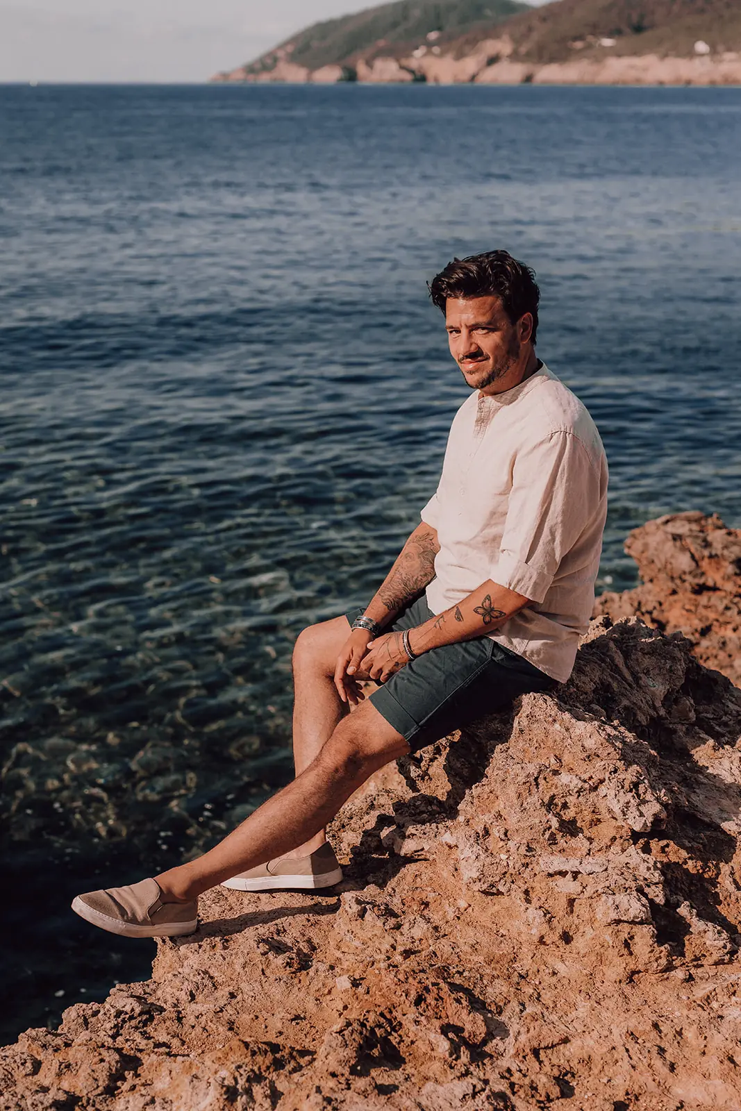 Rocco zit glimlachend op een rots boven het heldere zeewater